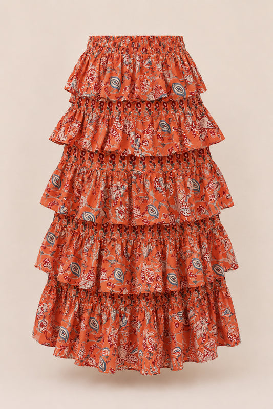 Model in handcrafted tiered maxi skirt standing among pink flowers under a blue sky.