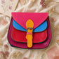 Handmade Leather Crossbody Bag in vibrant fuchsia, blue, and red colors, featuring a stylish buckle detail on a floral background.