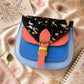 Handmade Leather Crossbody Bag - Romy - Painted Garden - Stardust displayed on a soft fabric with flowers and sketches.