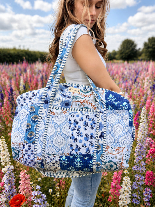 Model carrying a handmade patchwork duffle bag in a colorful flower field under a bright blue sky.