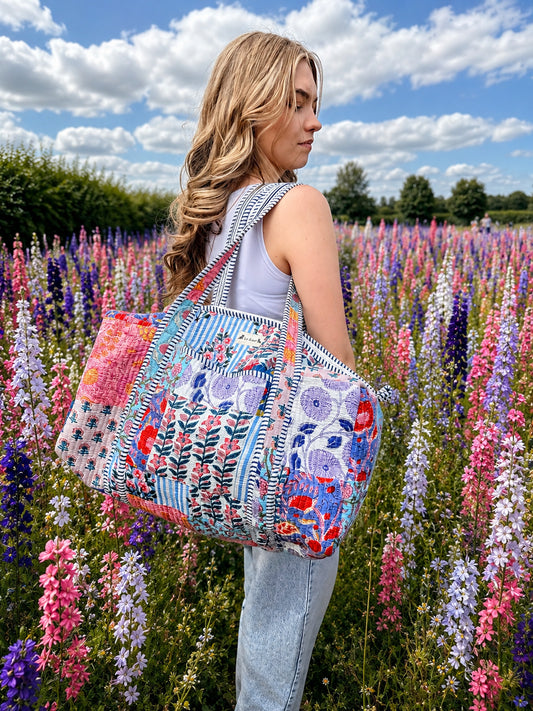Model carrying a handmade patchwork duffle bag in a colorful flower field under a bright blue sky.